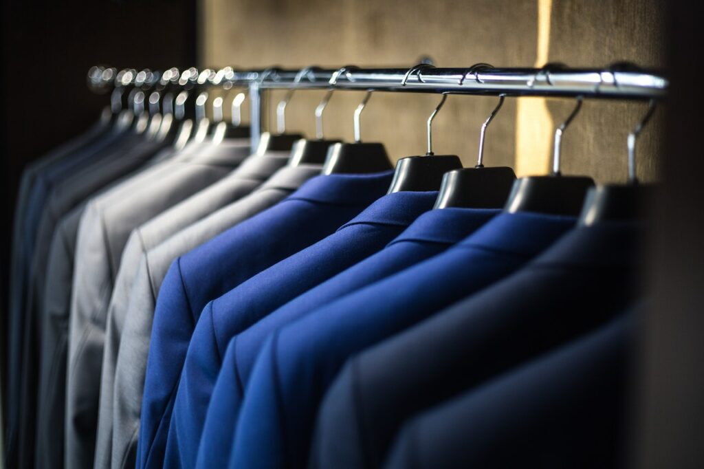 Blue, grey, and black suits hanging on hangers on a metal pipe.