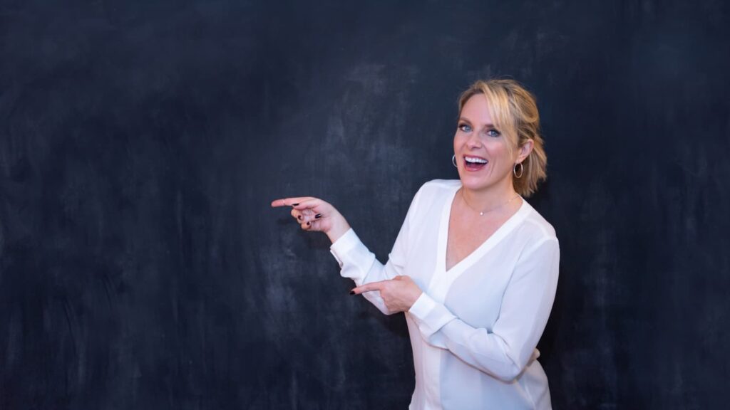 Kerry Barrett standing in front of a black board wearing a white shirt, smiling and pointing to the left.