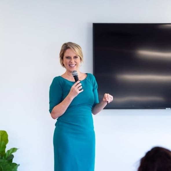 Kerry Barrett at a speaking engagement event holding a microphone and smiling. She is wearing a teal dress.