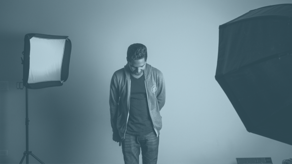 A man wearing a jacket and standing between pieces of lighting equipment, looking down at his feet