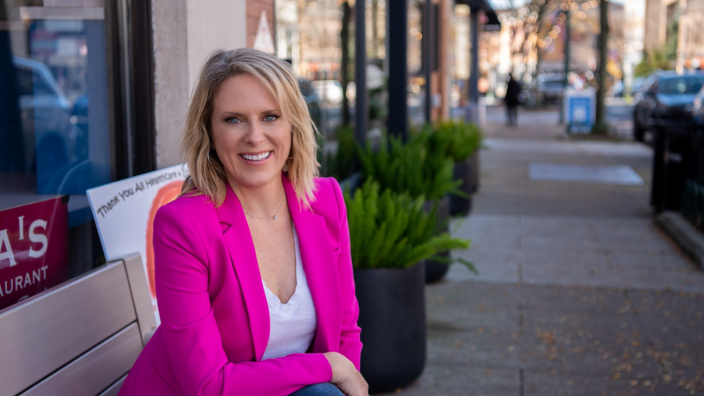 A white woman sitting on an outdoor bench smiling at the camera; she is wearing a brightly colored blazer