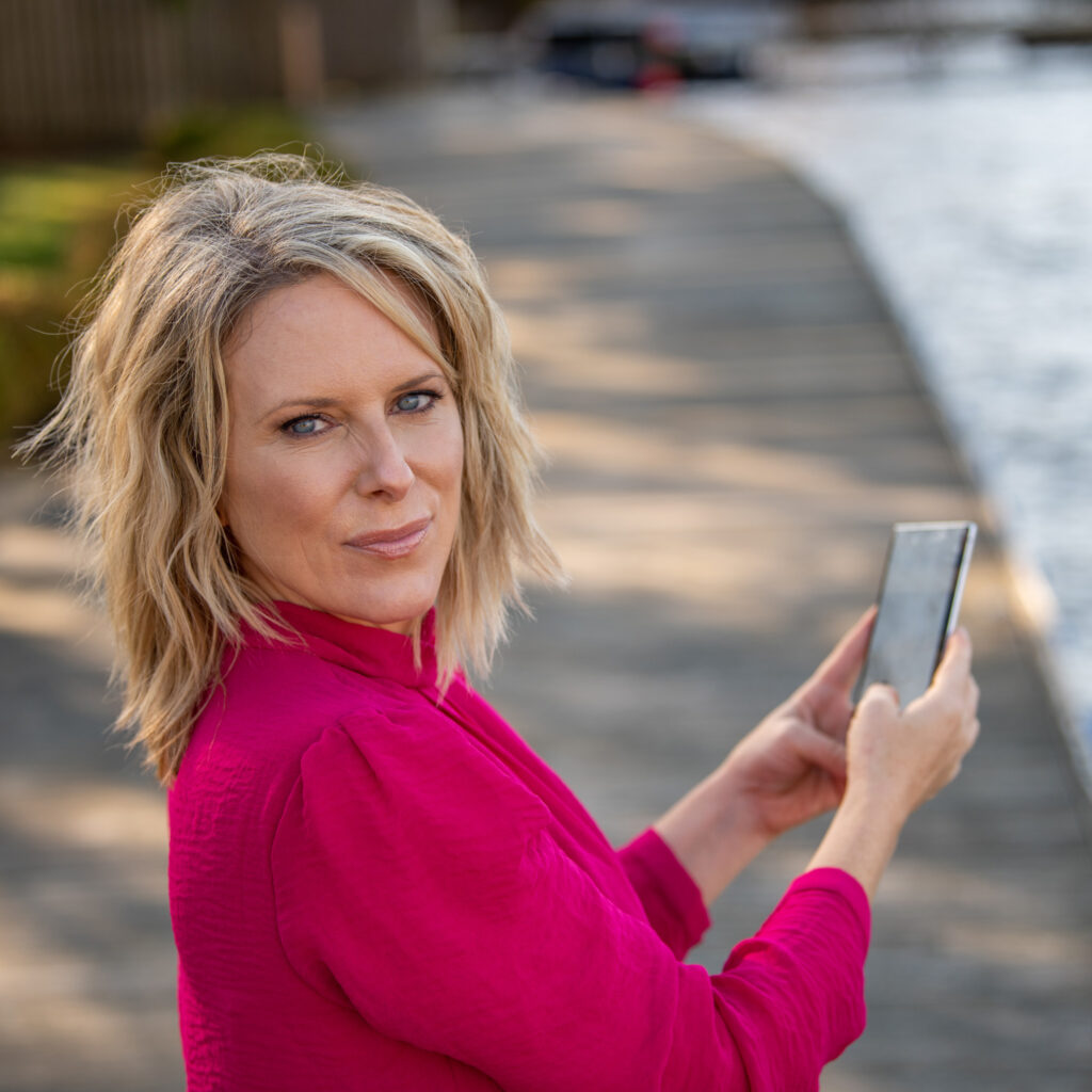 A blonde woman (Kerry Barrett) holding up a phone and looking over her right shoulder toward the camera.